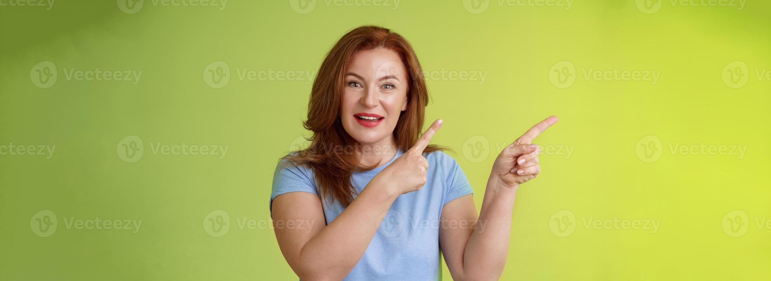 Curious enthusiastic gorgeous redhead female intrigued pointing upper left corner asking question interesting product talk shop assistant consulting promo stand green background blue t-shirt photo