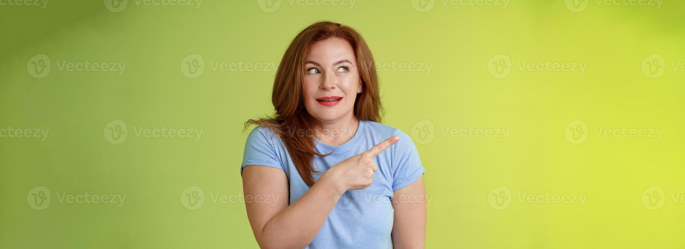 Excited intrigued middle-aged mature redhead woman pointing gazing left side copy space curiously smiling thrilled like interesting promo willing check-out great advertisement green background photo