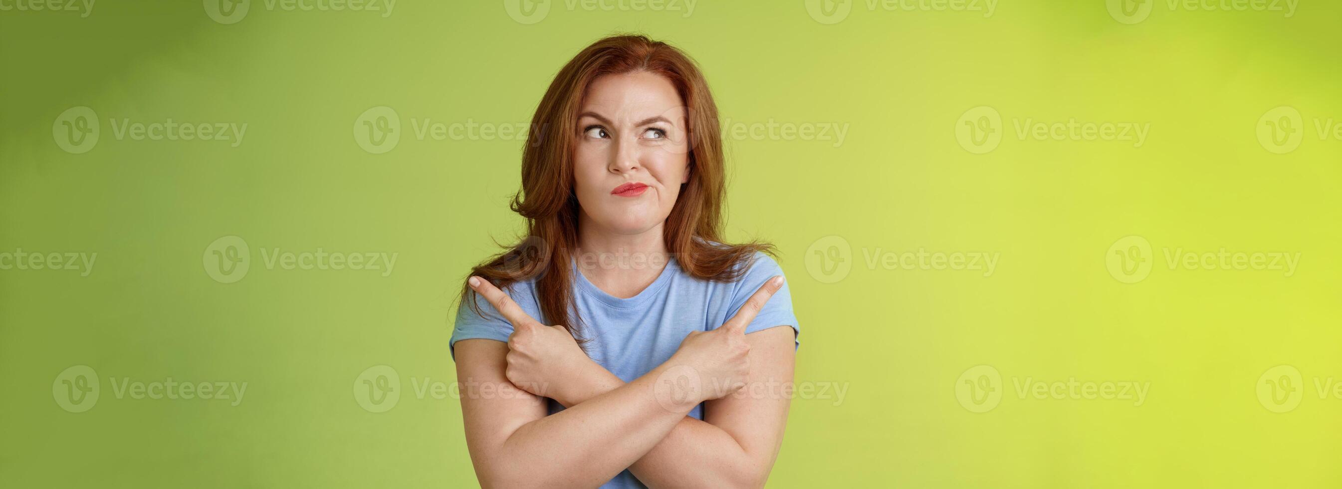 Time make right choice. Perplexed hesitant redhead middle-aged female customer deciding between few variants cross arms body pointing sideways look left smirking unsure doubting choosing product photo