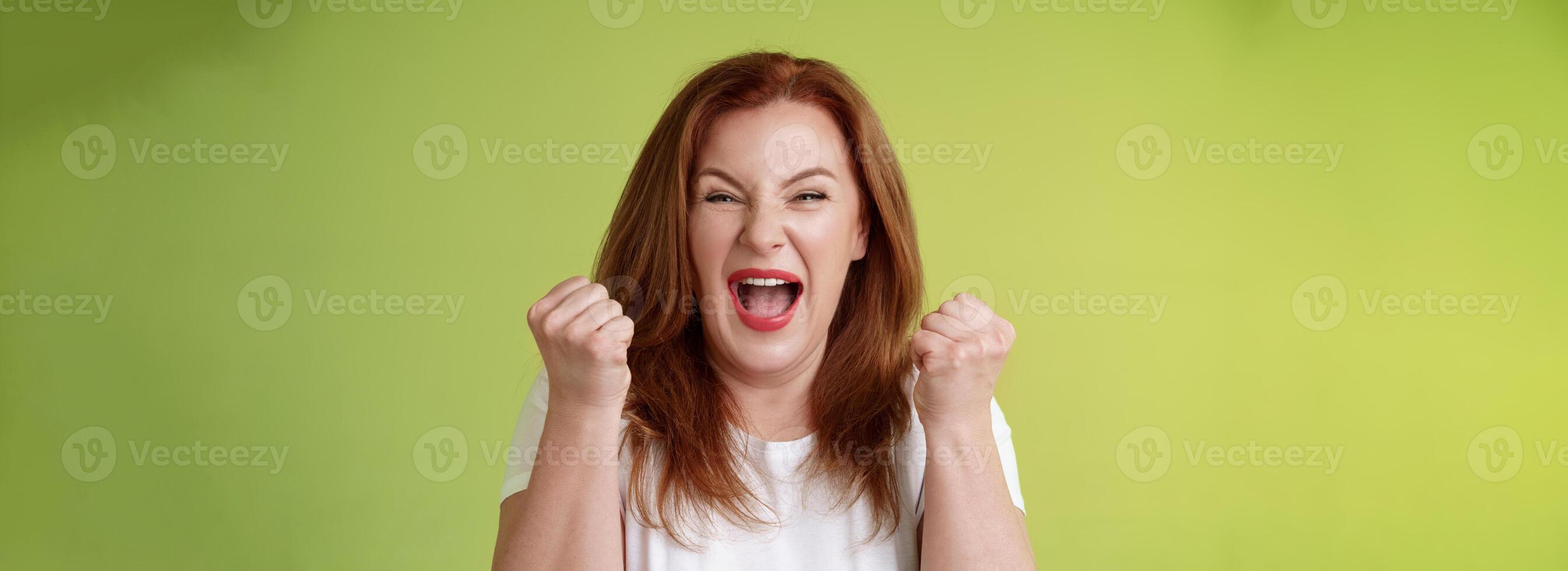 Yeah we did it. Joyful lucky redhead middle-aged female winner pump fists up celebration success gesture yelling triumph joy smiling broadly celebrate awesome news stand green background happy photo