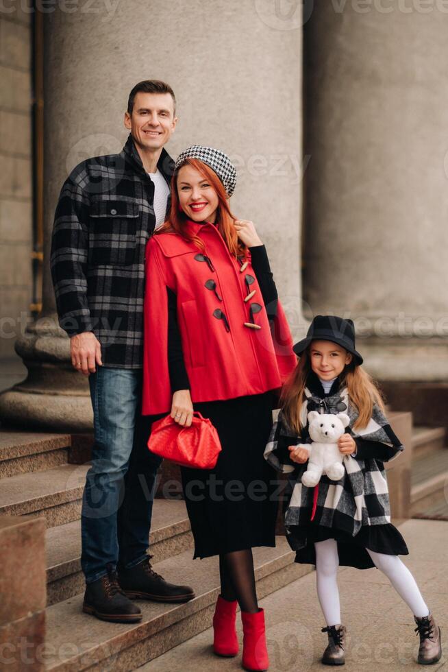 un elegante familia de Tres es en pueblo en el caer, y el niña con el relleno juguete es alegre foto