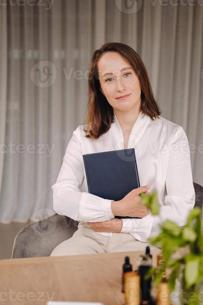 portrait of a smiling girl-woman sitting in an armchair. An aromatherapist in a white blouse is sitting in the office photo