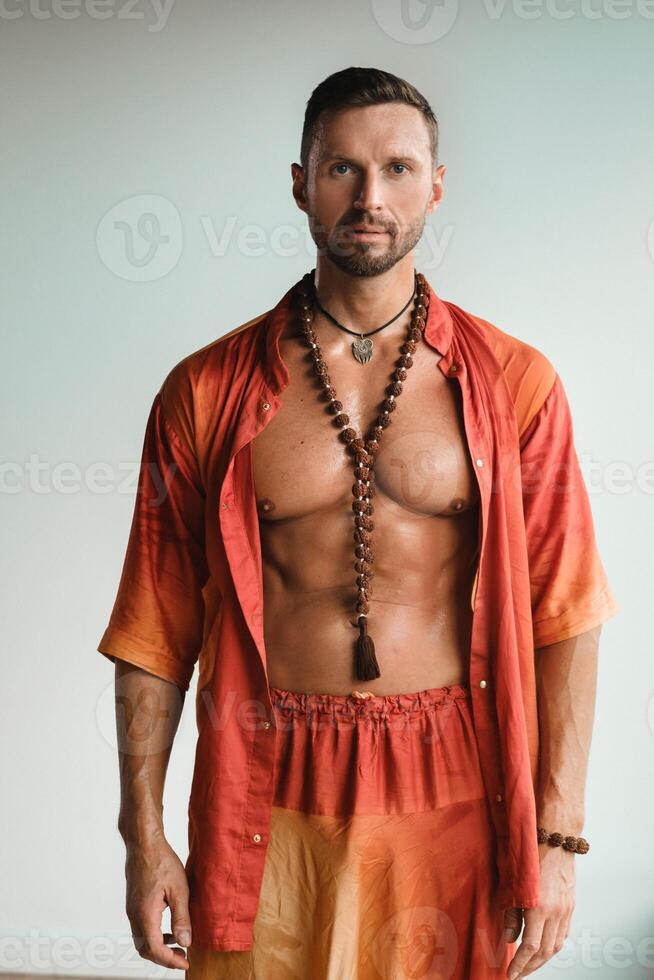 A muscular man with a naked torso in orange clothes stands in a room on a light background photo