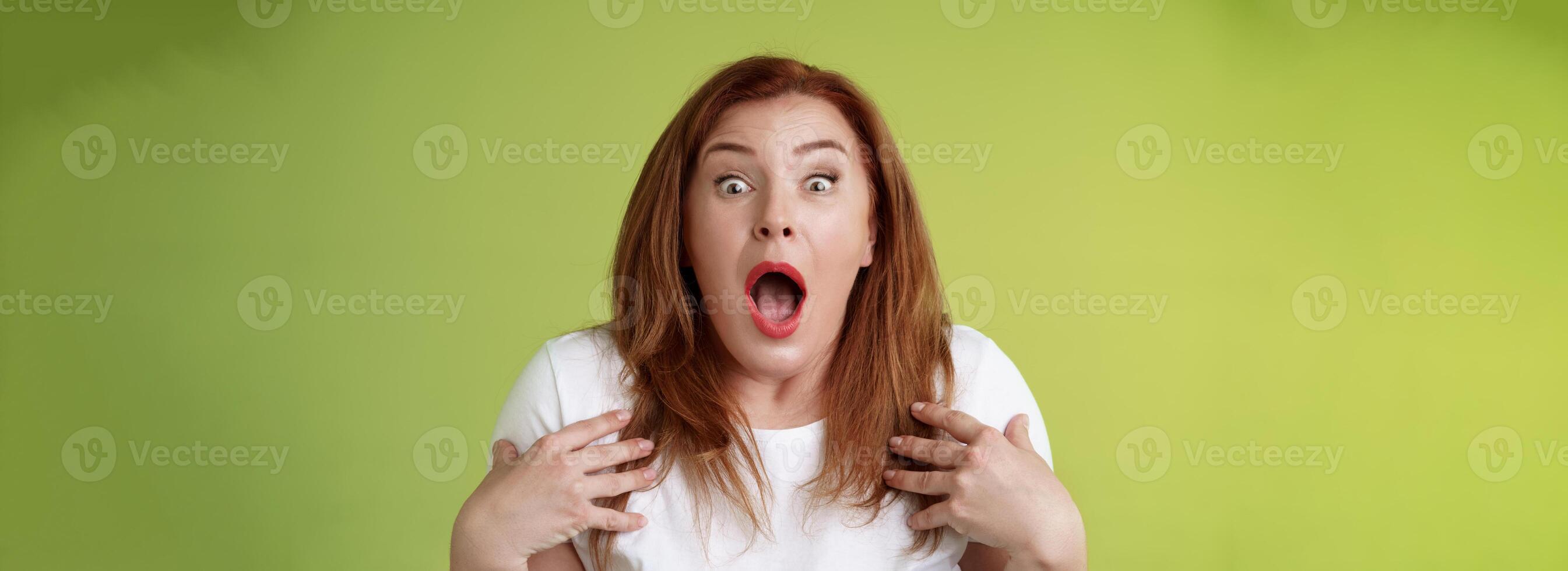 Shocked panicking redhead middle-aged woman gasping drop jaw open mouth stare camera freak-out anxious pointing herself impressed terrified frustrated nervously react green background photo