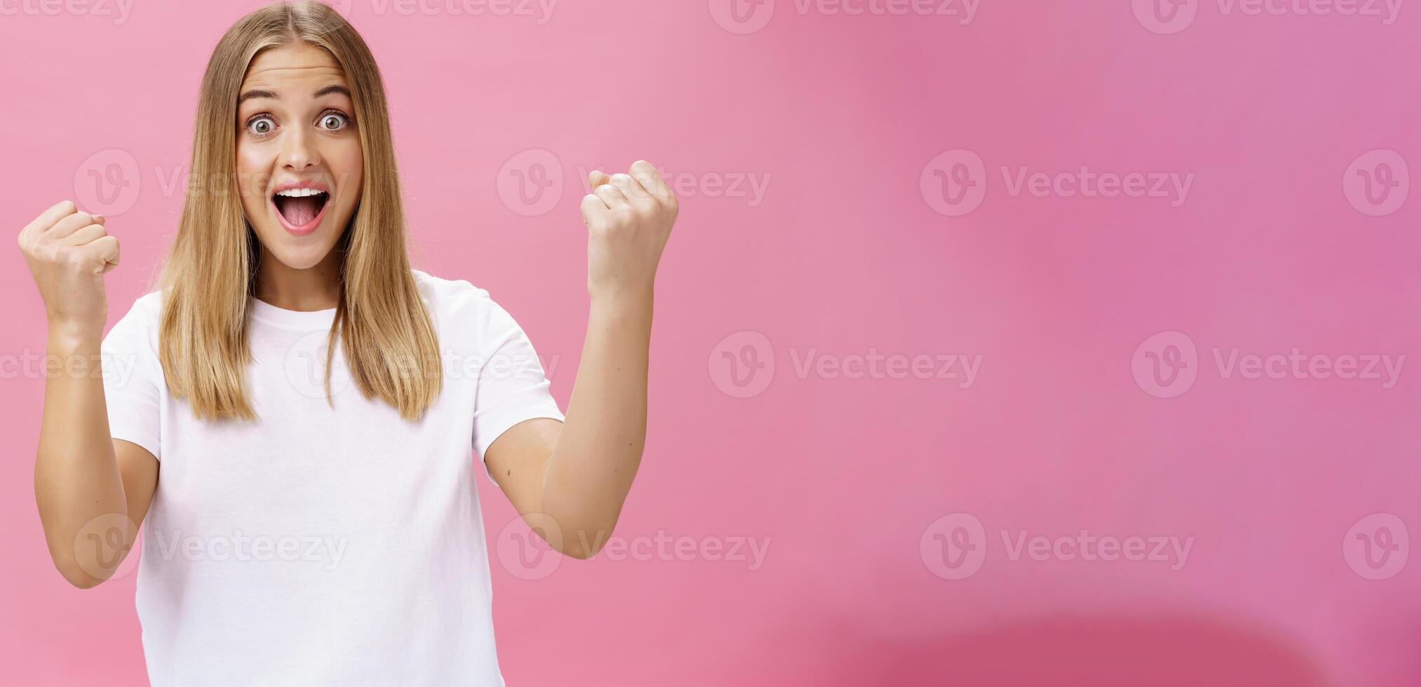 emocionado alegre y optimista encantador mujer con justa pelo en blanco camiseta levantamiento puños en victoria y triunfo Gritando si en éxito y asombro en pie apoyo en contra rosado antecedentes foto