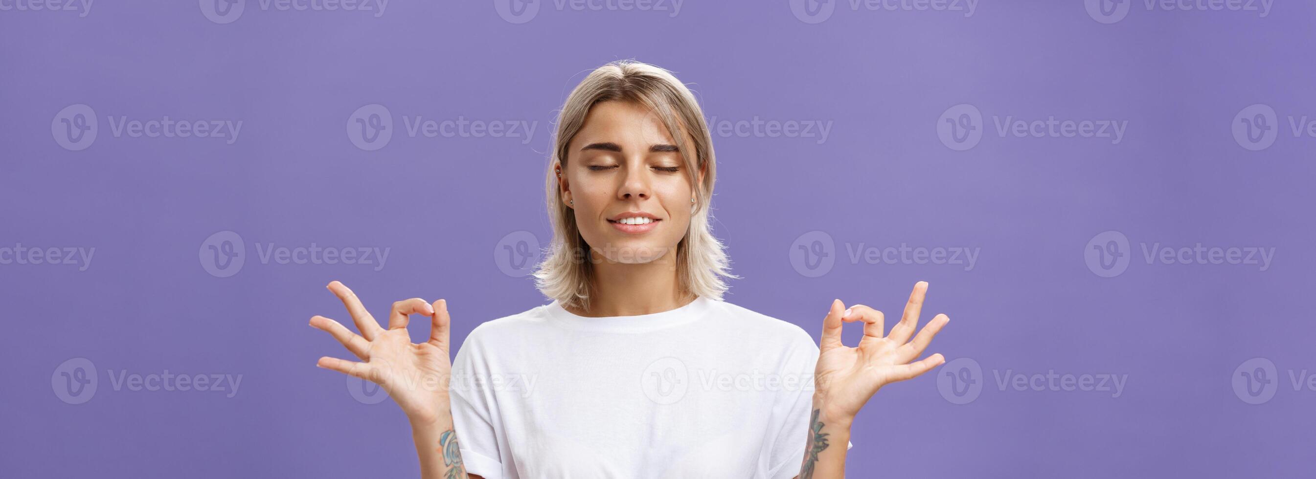 Peaceful calm good-looking female blonde in white t-shirt closing eyes smiling relieved and happy feeling satisfied with life standing in lotus pose with zen gesture over purple background photo