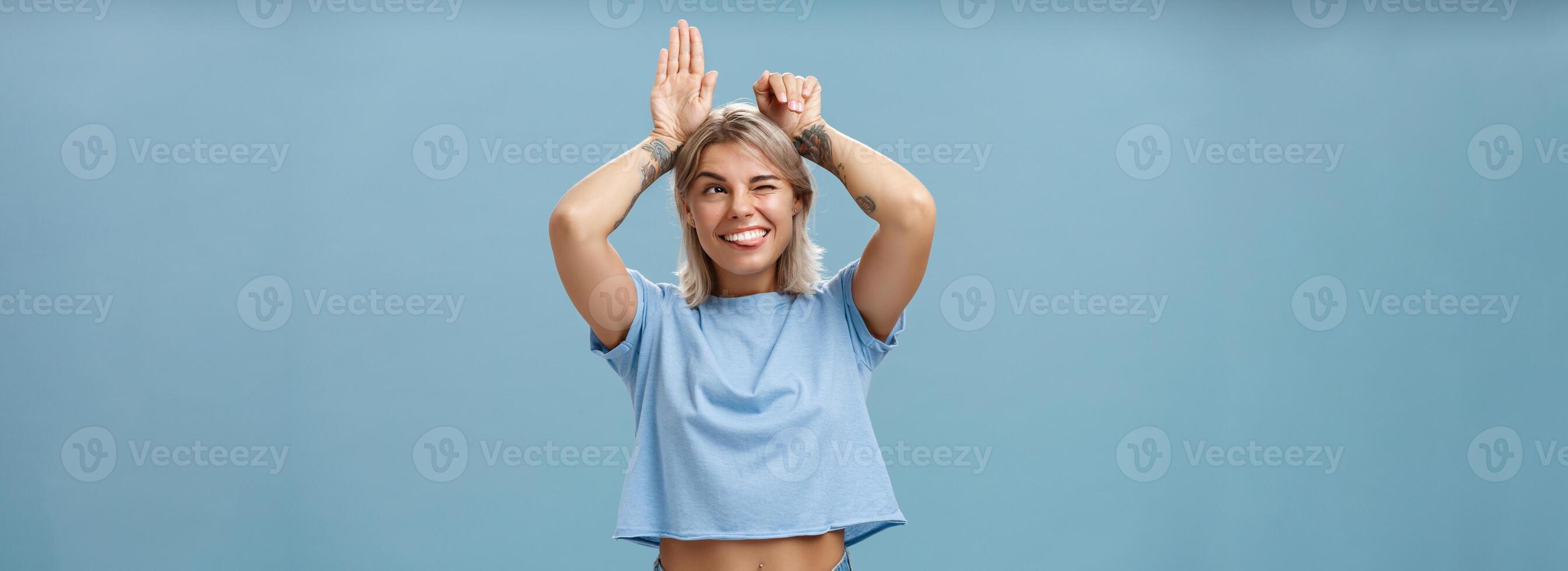 Studio shot of entertained carefree and emotive happy charming woman with tattoos on arms acting like bunny with palms on head winking joyfully smiling and sticking out tongue over blue wall photo