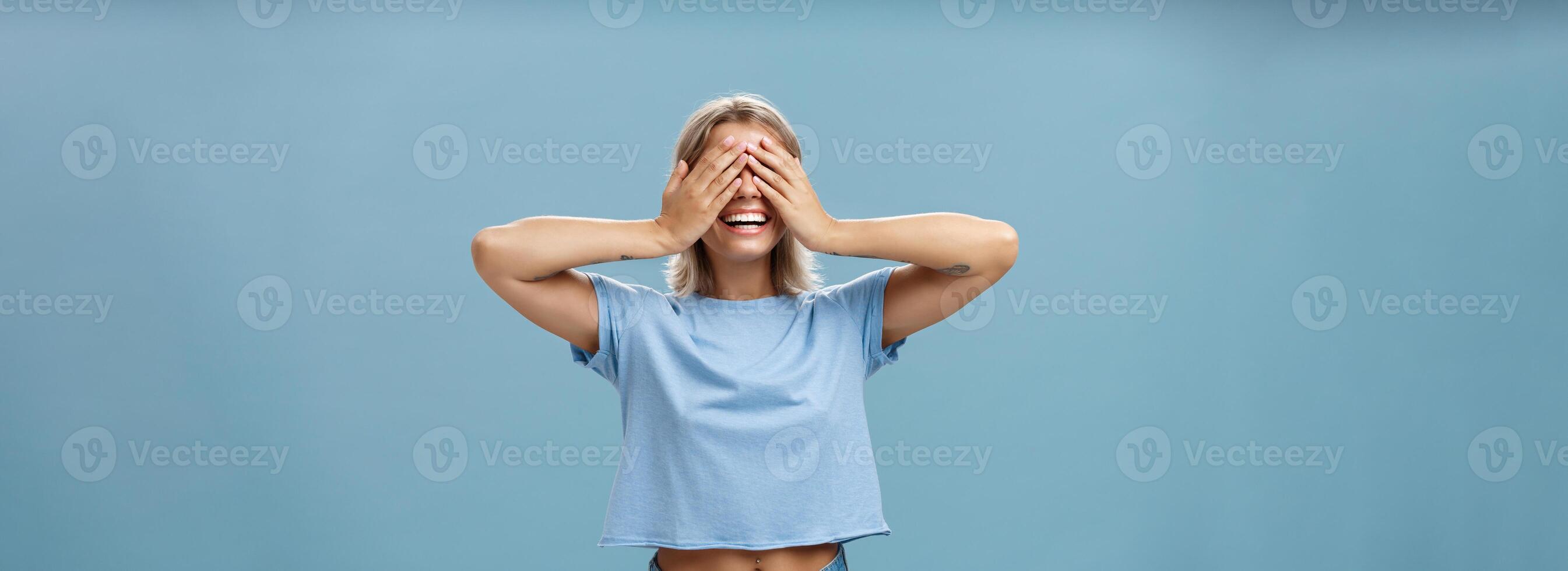 Hide if you can. Portrait of carefree and happy good-looking playful girl with blonde hair smiling broadly while counting ten and playing hide-and-seek with closed eyes over palms near blue wall photo