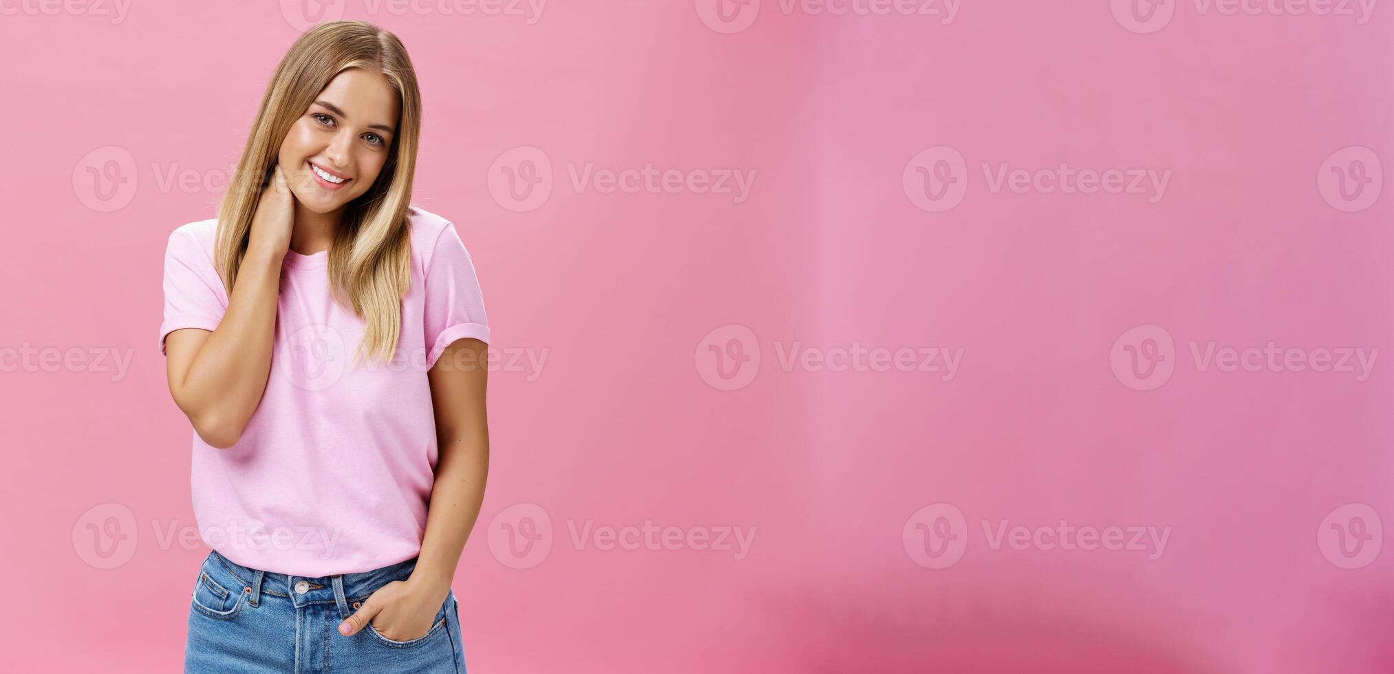 Portrait of shy and timid attractive european woman with fair hair in casual outfit touching neck silly tilting head and smiling cute at camera standing over pink background photo