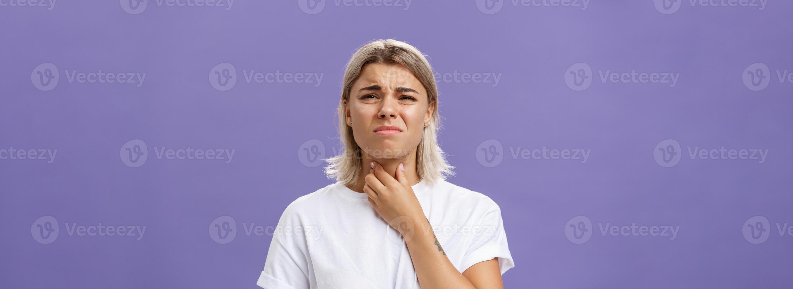 Displeased young stylish caucasian woman with blond hair and tanned skin feeling discomfort in throat having inflammation or feeling cold touching neck and frowning dissatisfied from pain photo