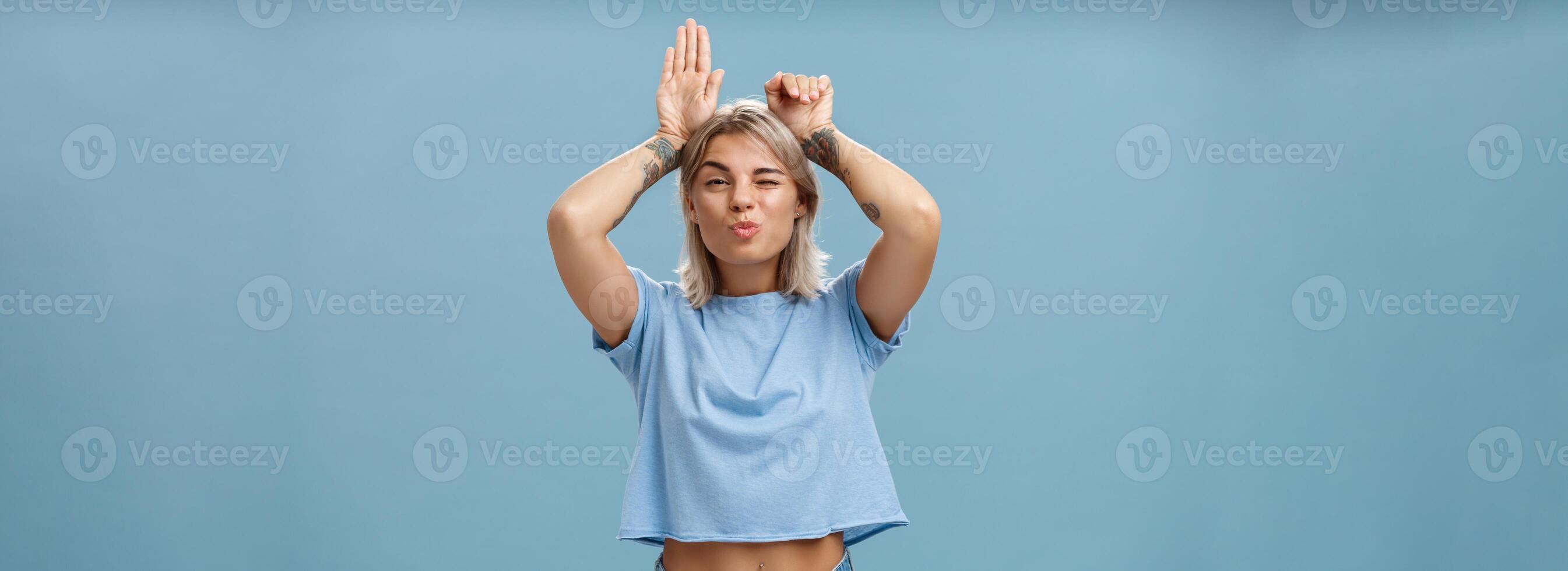 Flirty attractive and stylish cool girlfriend with tattooed arms and pierced belly winking folding lips in air kiss showing bunny ears with palms on head standing over blue background playful photo