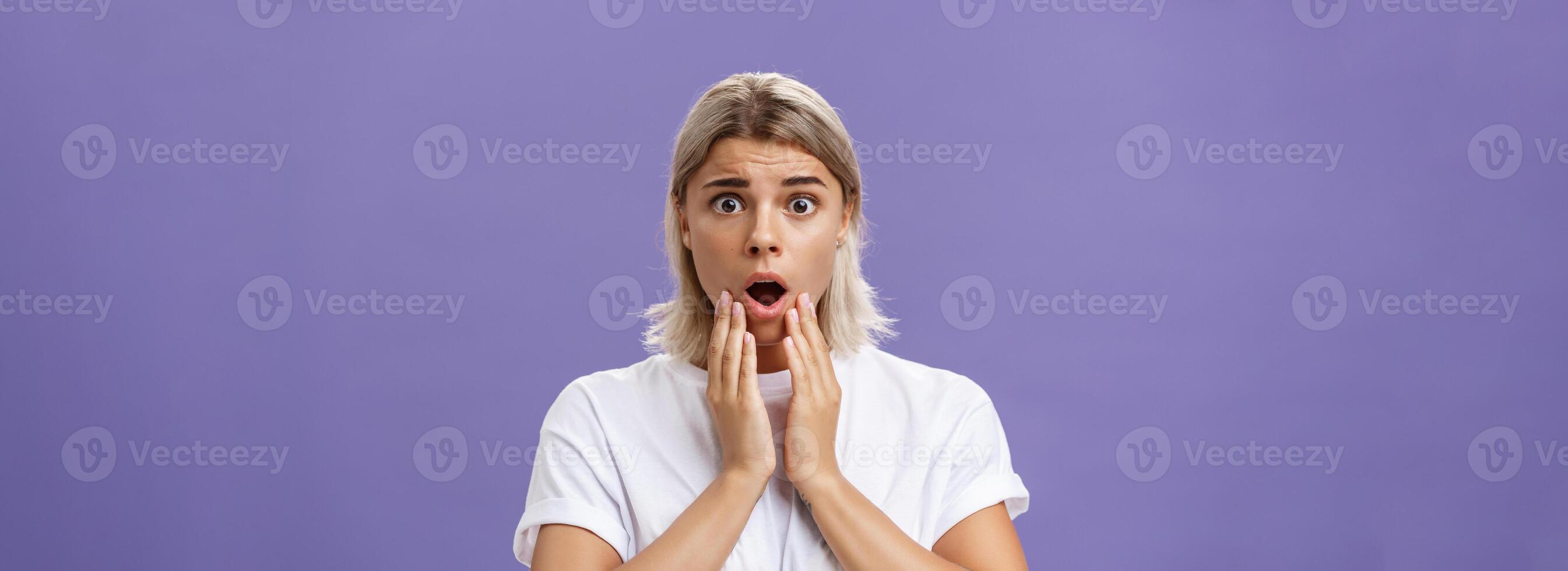 Waist-up shot of worried and shocked woman expressing empathy frowning holding hands near opened mouth feeling sorry and nervous hearing terrible disaster happened standing over purple wall photo