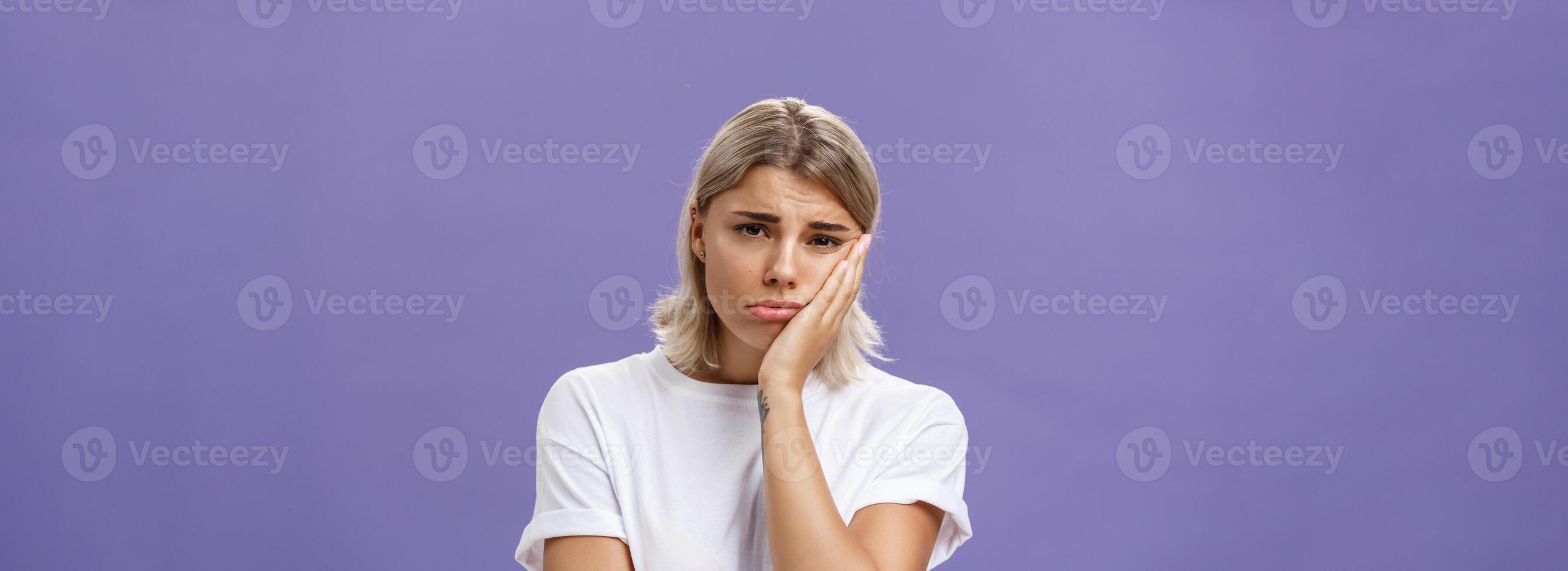 Waist-up shot of unhappy miserable and sad cute blonde female in white casual t-shirt pursing lips leaning face on palm and frowning from disappointement and regret over purple background photo