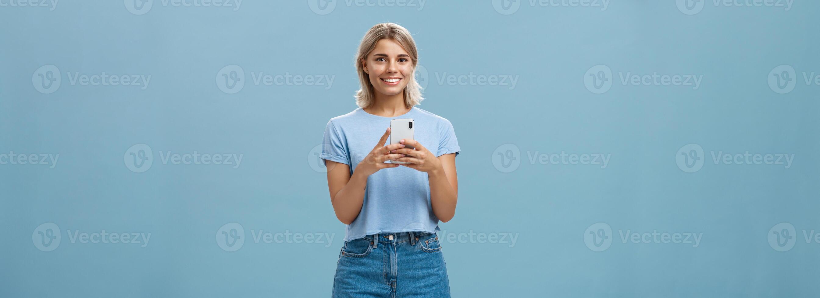 nuevo teléfono es fantástico. retrato de satisfecho contento joven moderno rubio mujer en de moda atuendo con rubio corto Corte de pelo sonriente alegre desde deleite participación teléfono inteligente cerca cofre mensajes de texto o utilizando aplicaciones foto