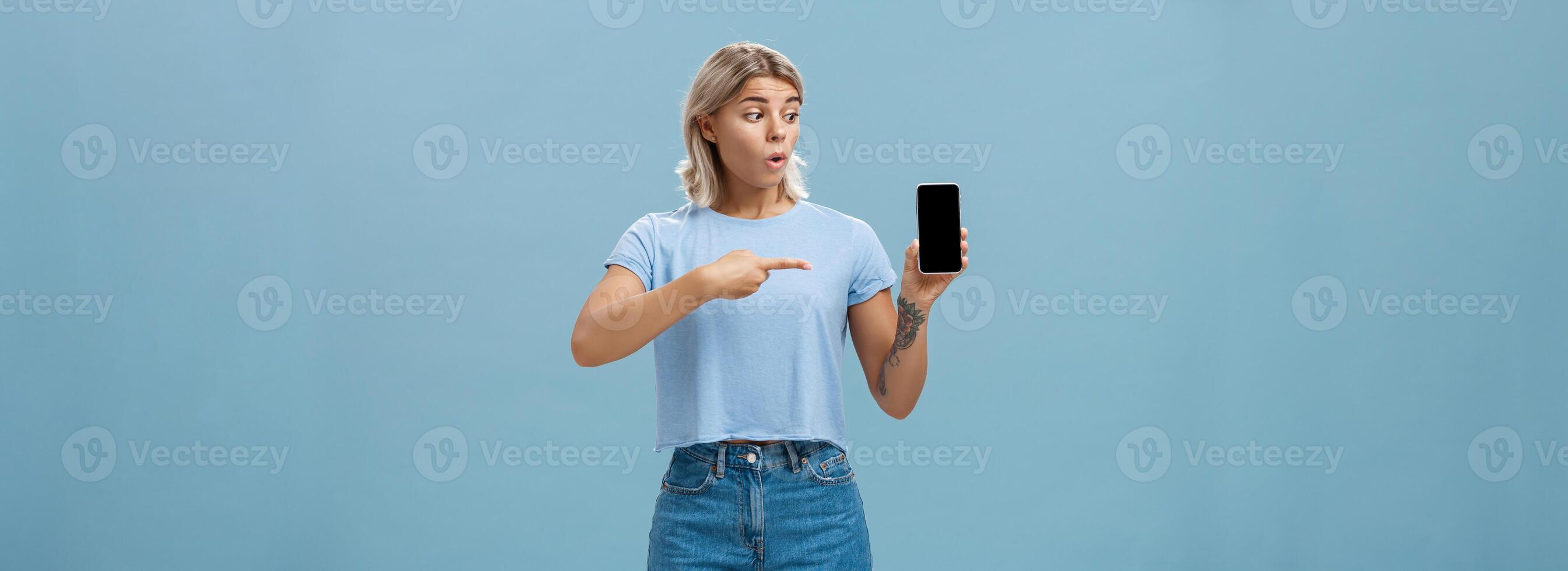 You should download app. Portrait of amazed shocked attractive blonde girl in trendy summer t-shirt holding smartphone gasping from surprise pointing at gadget screen posing over blue background photo