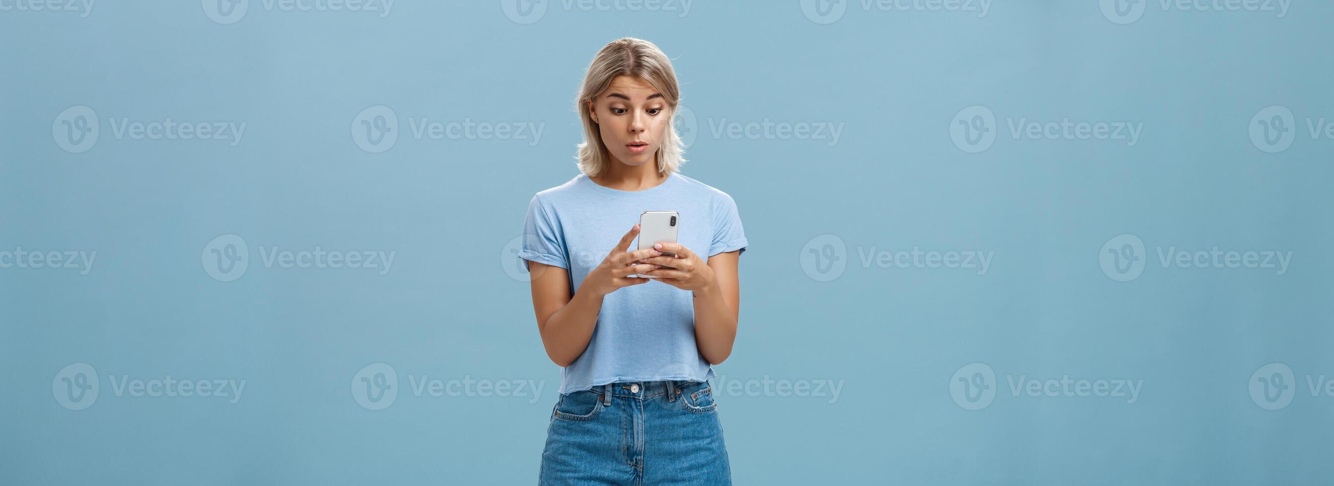 Indoor shot of surprised young caucasian girl receiving unexpected invitation via smartphone reading strange message in cellphone staring stunned and confused at screen over blue background photo