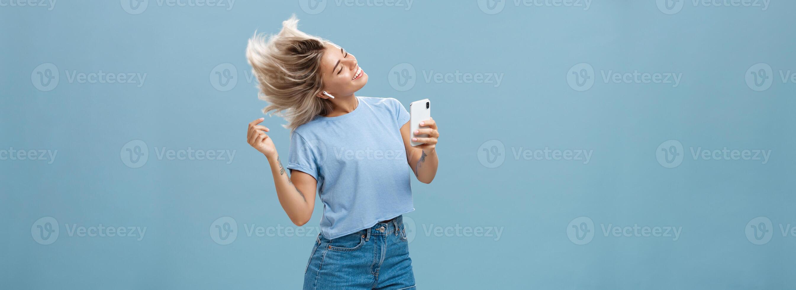 Girl enjoying cool bits in brand new wireless earphones advertising earbuds in own blog recodring video via smartphone dancing from joy and delight smiling listening music over blue background photo