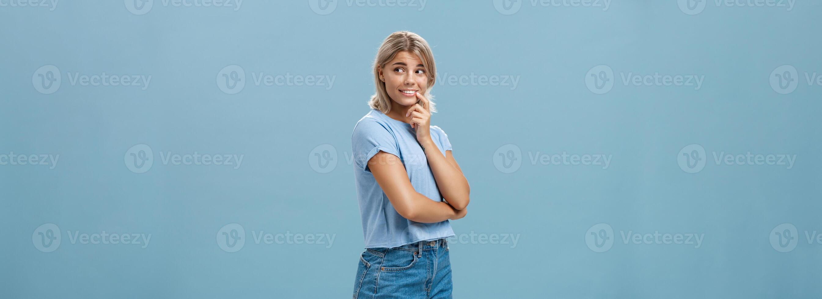 Indoor shot of feminine romantic and timid good-looking european female with fair hair holding finger on lip smiling curious and intrigued while turning right and observing interesting thing photo