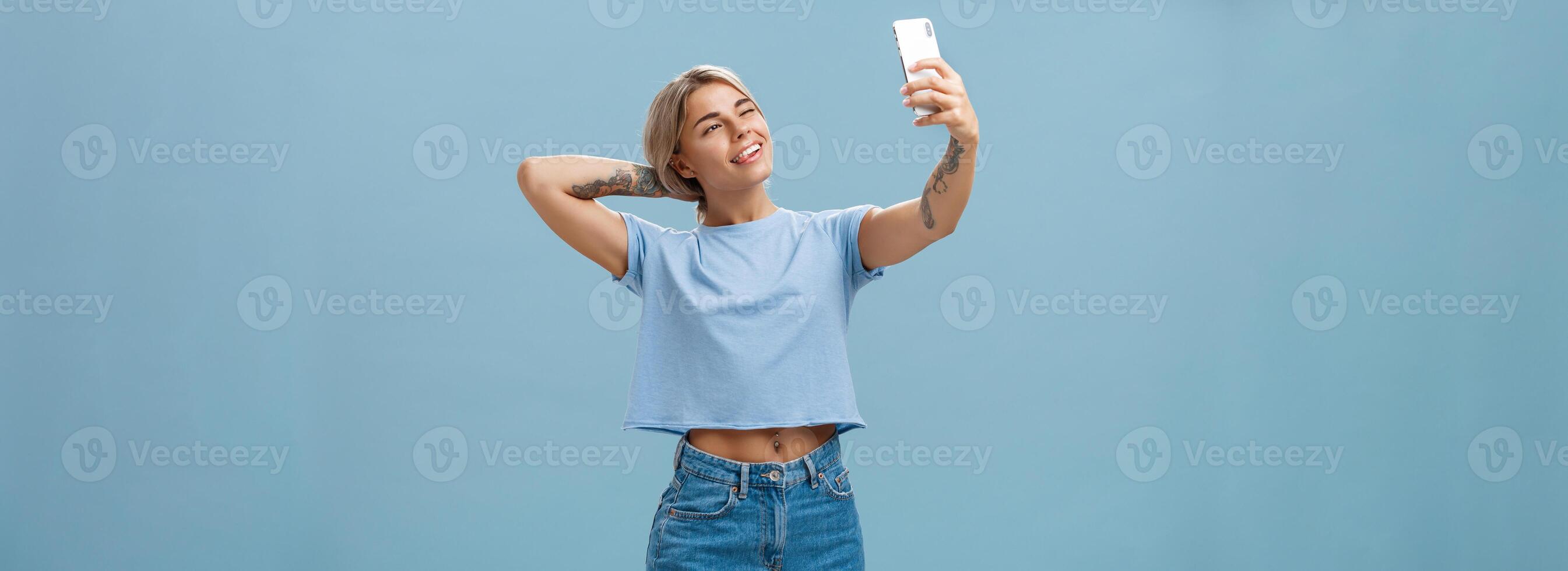 nuevo enviar en mi Blog. elegante femenino y sociable guapo joven hembra posando para selfie con tirado brazo y teléfono inteligente pega fuera lengua mientras haciendo caras a dispositivo pantalla terminado azul pared foto