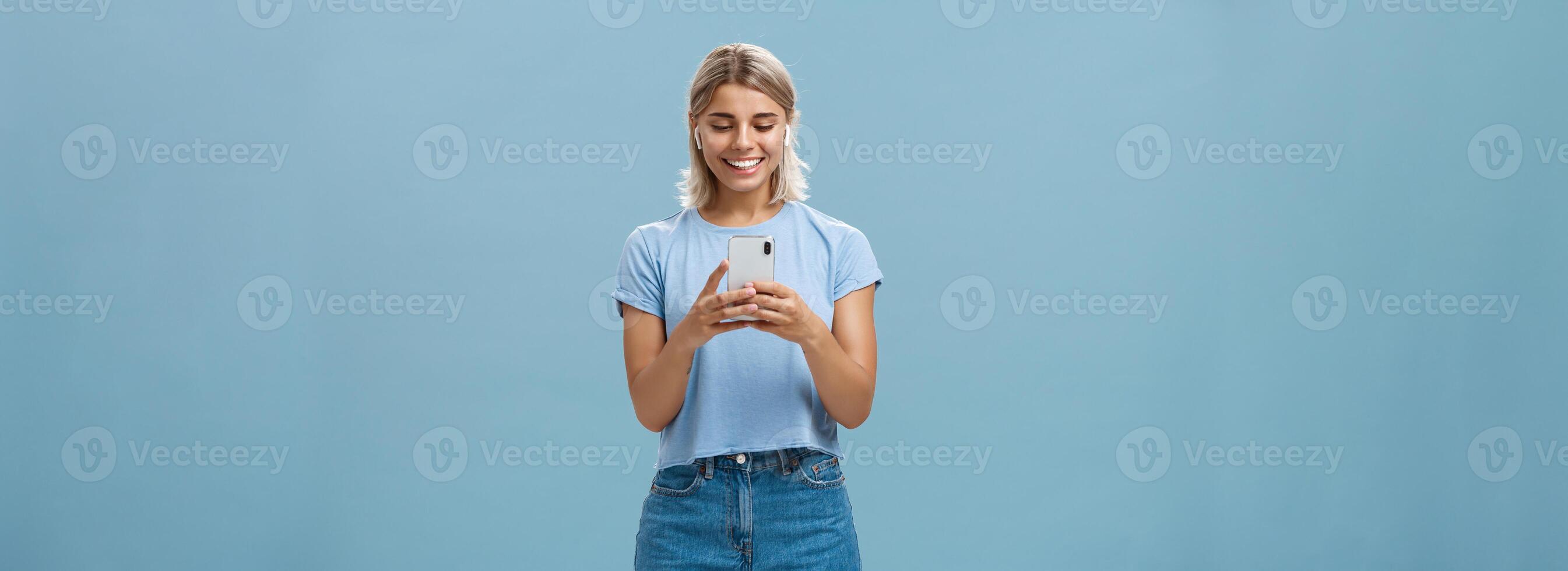 Girl having funny convesation, speaking with friend via video messages in wireless earphones holding smartphone in both hands smiling happily at device screen being entertained and amused photo