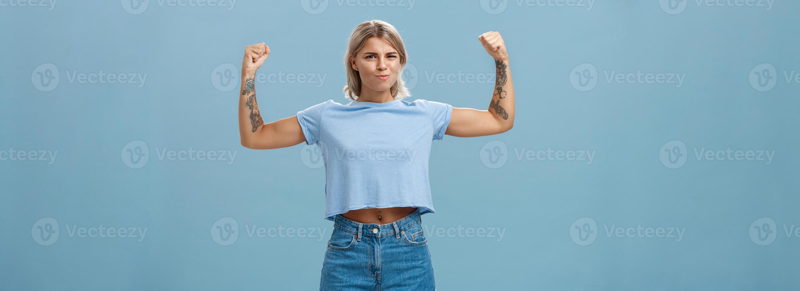 fuerte y poderoso guapo deportistas con tatuajes en camiseta y pantalones cortos levantamiento brazos demostración grande músculos y bíceps sonriente con orgullo mientras fanfarronería con físico fuerza terminado azul pared foto