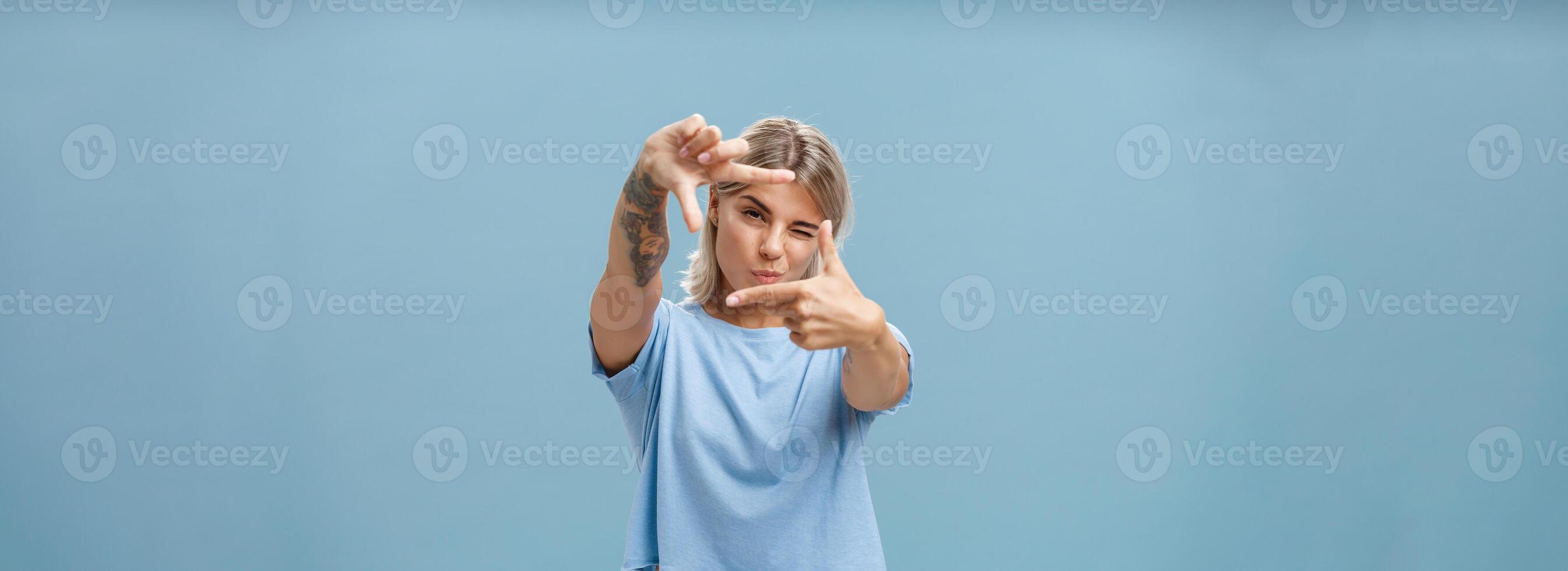 Artistic and creative female designer in stylish t-shirt closing one eye folding lip and making frame gesture while looking through it as if taking measurement or picturing something over blue wall photo