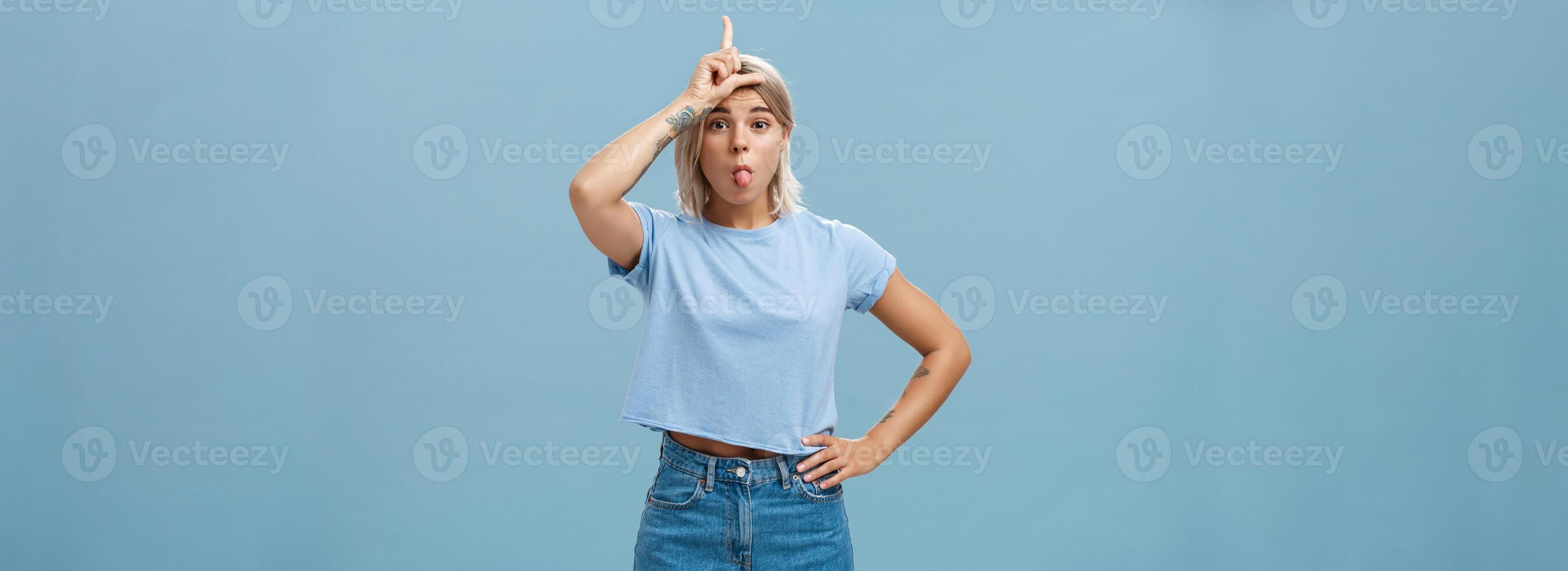 estudio Disparo de juguetón triunfando guapo rubia hembra con tatuado brazos demostración letra l en frente y pega fuera lengua burlón terminado perdedor y celebrando propio victoria terminado azul pared foto