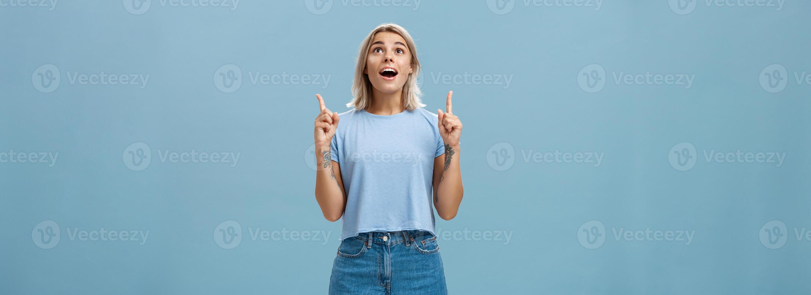 Indoor shot of impressed speechless attractive fair-haired female student in casual t-shirt and denim shorts dropping jaw from amazement pointing and looking up intrigued over blue wall photo