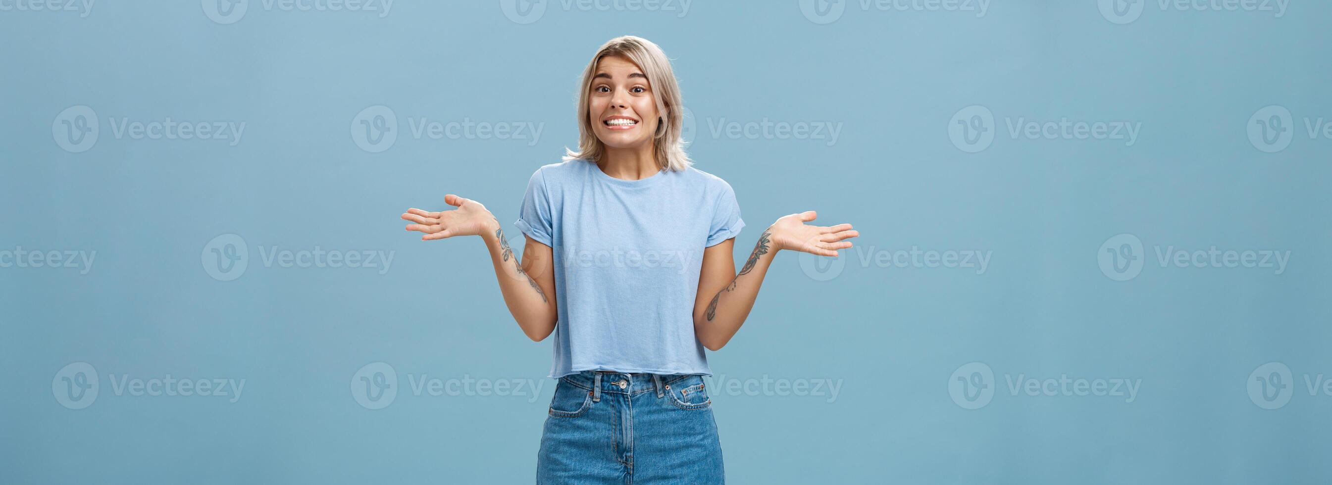 Uy yo hacer no saber. tonto linda europeo niña con justa pelo y frio tatuajes encogiéndose de hombros con palmas untado aparte en No idea gesto sonriente con lo siento Mira siendo inconsciente y despistado terminado azul pared foto