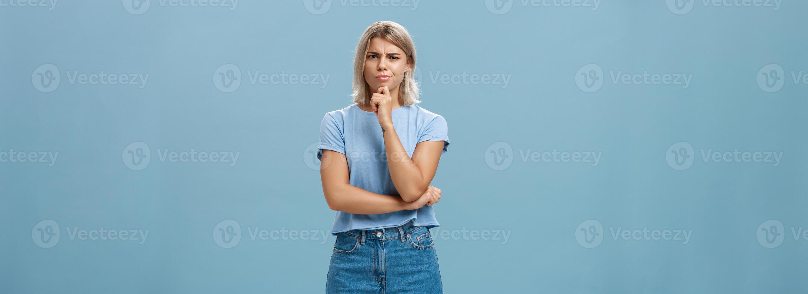 I doubt it good idea. Suspicious intense creative young female coworker in outdoor outfit frowning from doubtful thoughts holding hand on chin while thinking expressing disbelief over blue wall photo