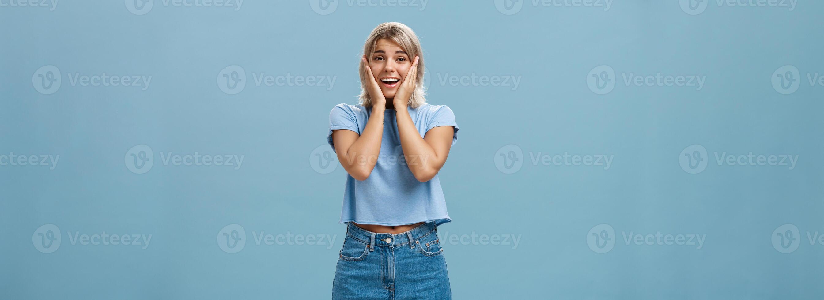 Oh my so cute. Portrait of touched tender good-looking athletic woman with fair hair holding palms on cheeks smiling being impressed and pleased standing in denim shorts over blue background photo