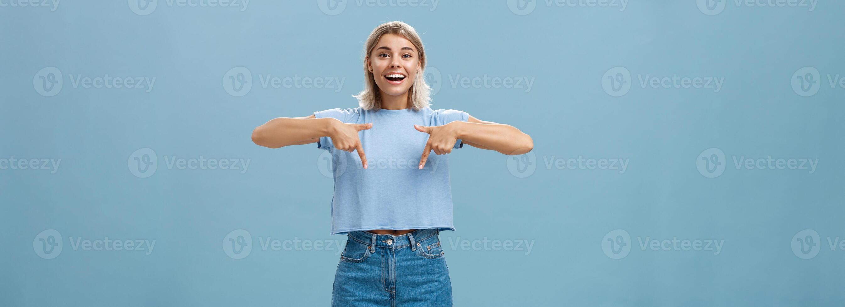 You have to see this. Portrait of joyful amused and happy stylish blonde female in trendy t-shirt pointing down with arms near chest smiling broadly showing amazing copy space over blue background photo