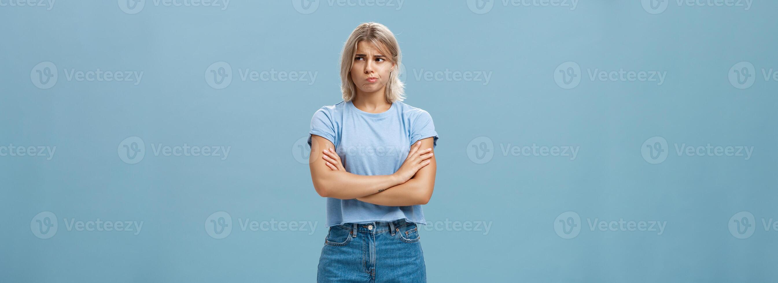 Uncertain troubled and perplexed attractive blond woman with tanned skin holding hands crossed on chest pouting and frowning looking right with worried unsure and doubtful look over blue wall photo
