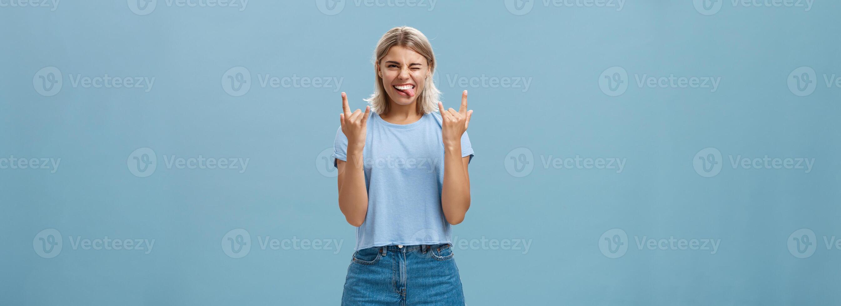 Let us rock this party. Portrait of joyful good-looking and carefree young artistic female musician with blond hair showing heavy metal gesture sticking out tongue and winking amused, having fun photo