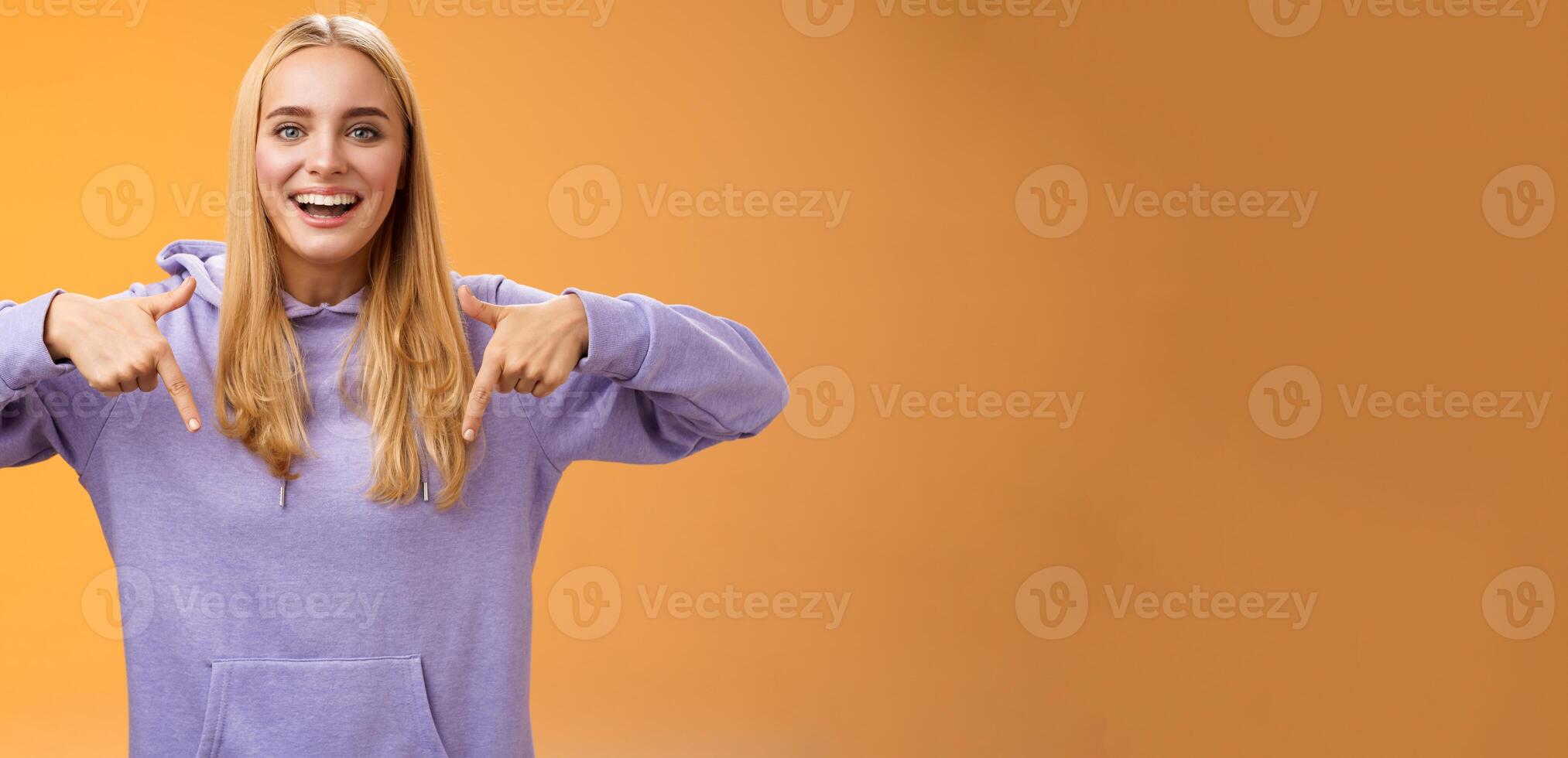Amused joyful nice blond girlfriend pointing down present cool new product smiling broadly recommending try check out standing orange background happily grinning in hoodie photo