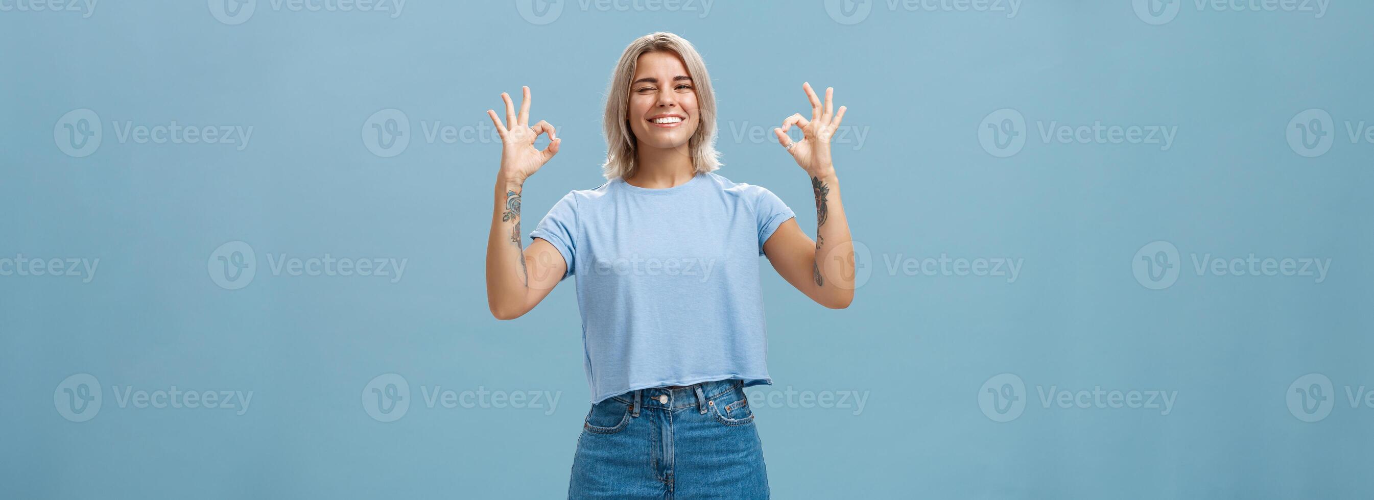 yo tiene usted cubierto. entusiasta atractivo y energizado hembra rubia con tatuado brazos demostración bueno o Perfecto firmar y parpadeo a asegurar amigo todo hecho sonriente alegremente terminado azul pared foto