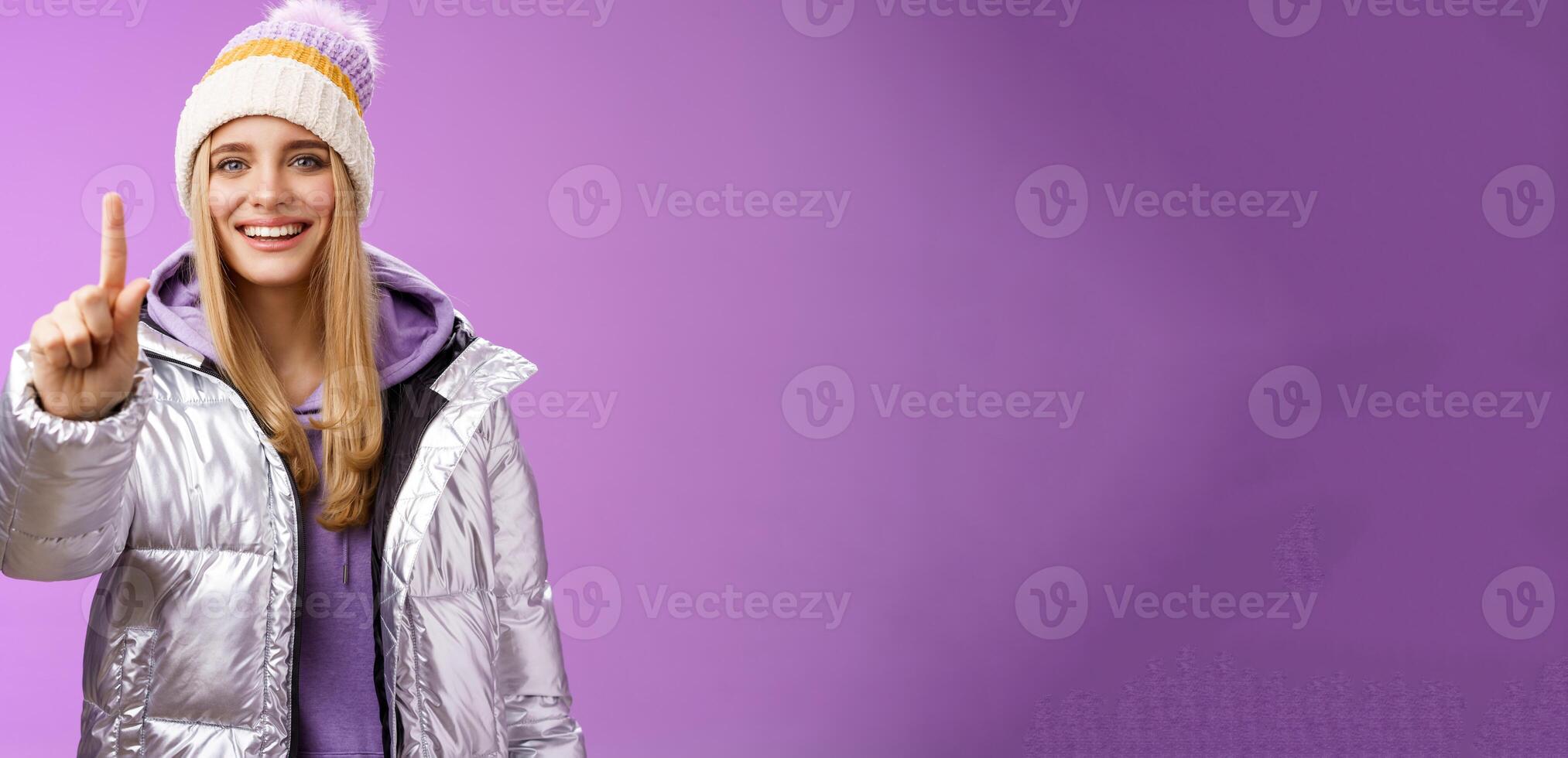 Excited cheerful fair-haired european girl in winter hat silver shiny jacket show number one index fingers give suggestion advice smiling broadly talking happily, purple background photo