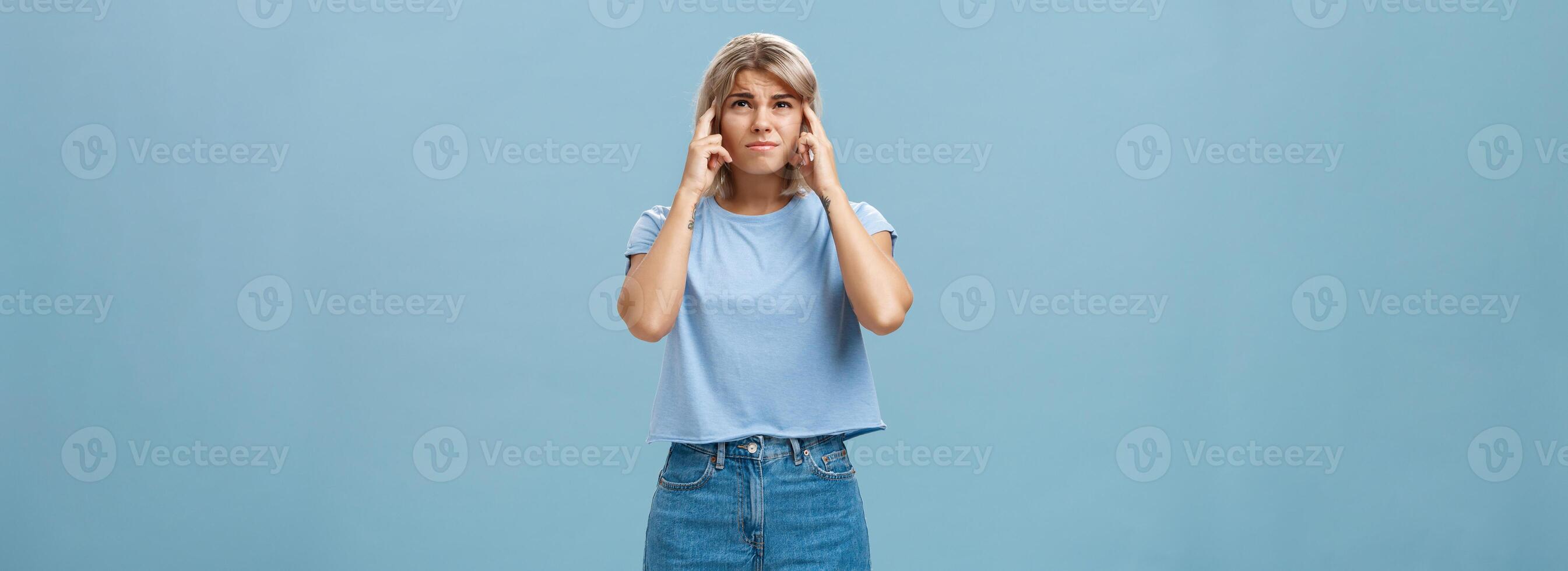 Girl cannot concentrate hearing loud noise comming from upstairs frowning feeling intense and dissatisfied closing ears with index fingers looking up while complaining on awful sound over blue wall photo