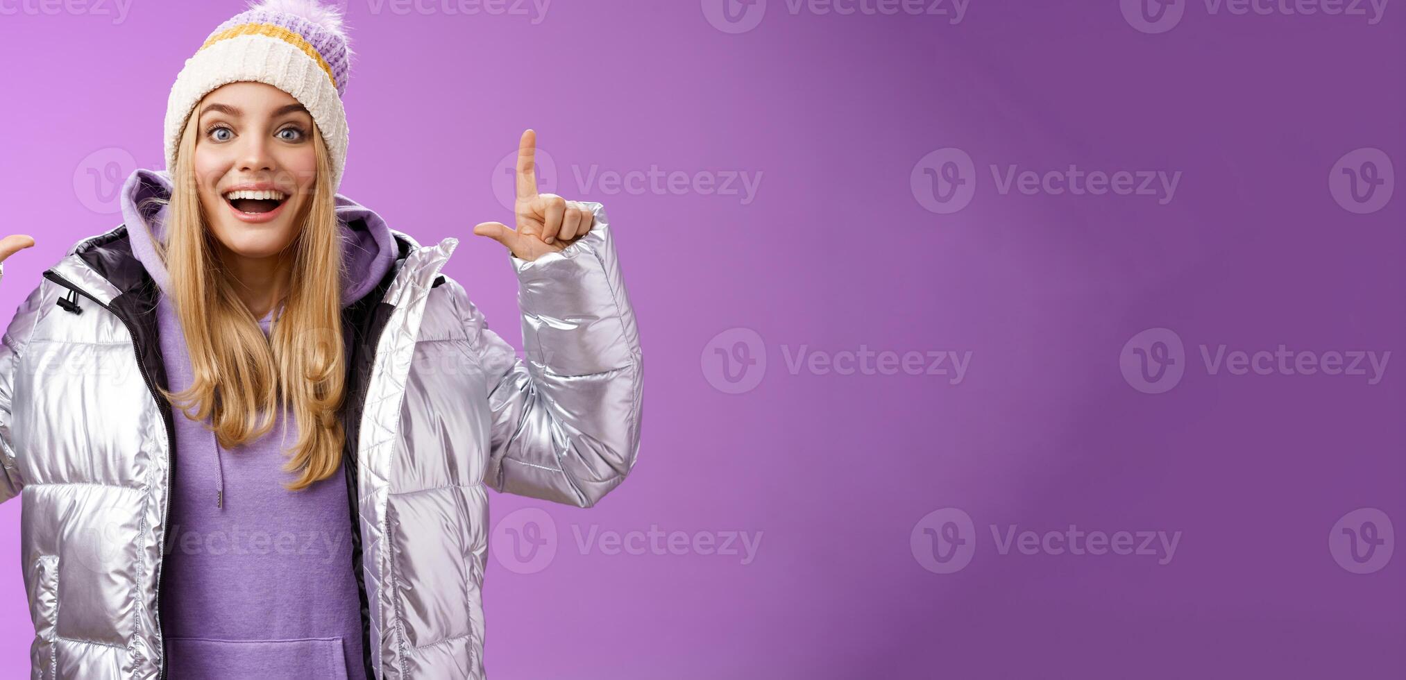 Excited carefree cheerful fair-haired european girl in silver jacket winter hat raising hands pointing up have excellent idea smiling broadly speaking passionately standing purple background photo