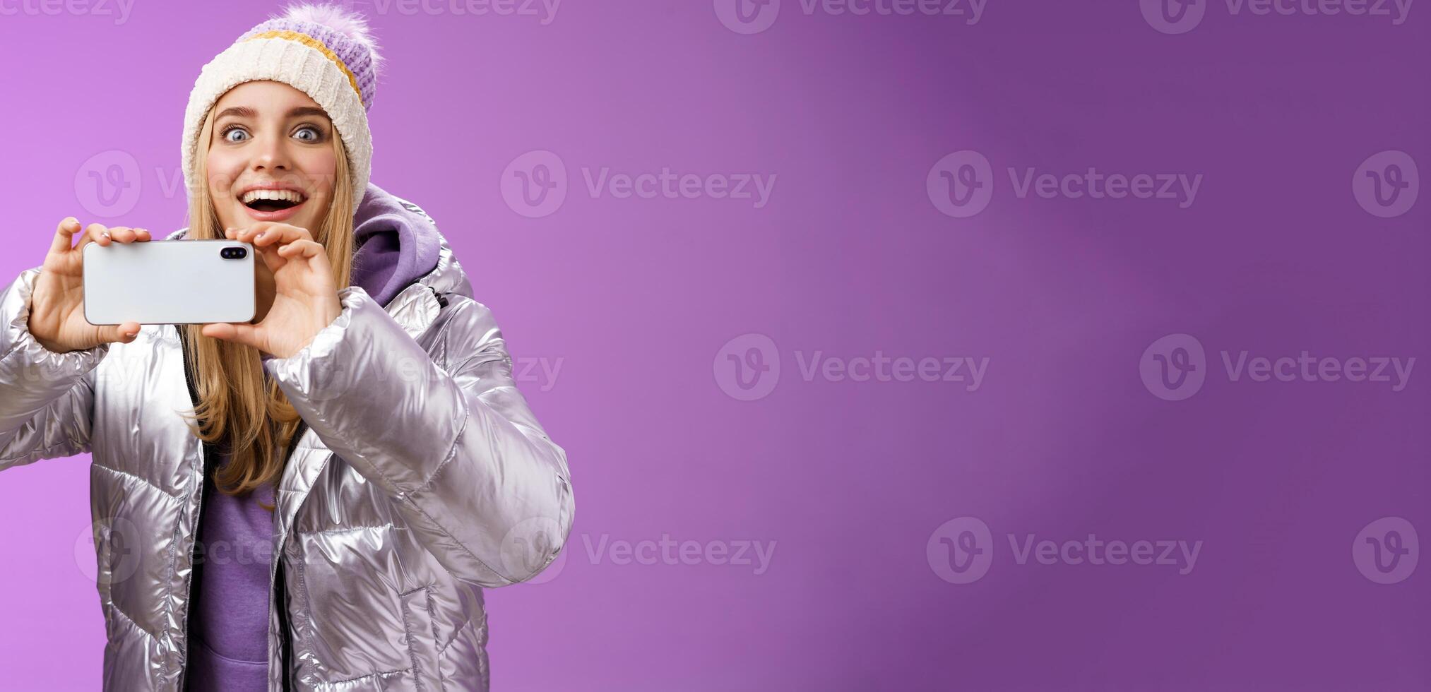 Amused fascinated female in silver jacket head smiling astonished excited look forward holding smartphone recording video taking shots famous person mobile phone camera, purple background photo