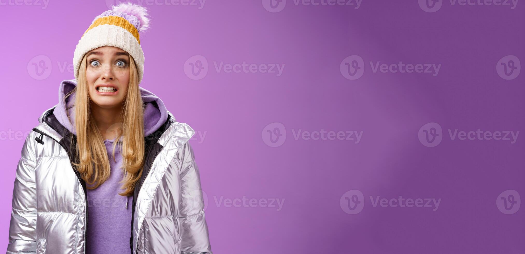 Awkward worried cute timid blond girl in silver jacket hoodie winter hat clench teeth popping eyes camera ooops make mistake standing nervous someone notice, purple background photo