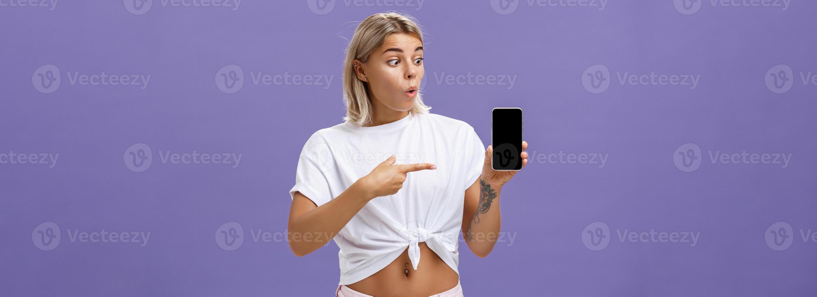 Waist-up shot of impressed and delighted gorgeous european female with blond hair and tattoos looking and pointing at awesome brand new smartphone standing over purple background photo