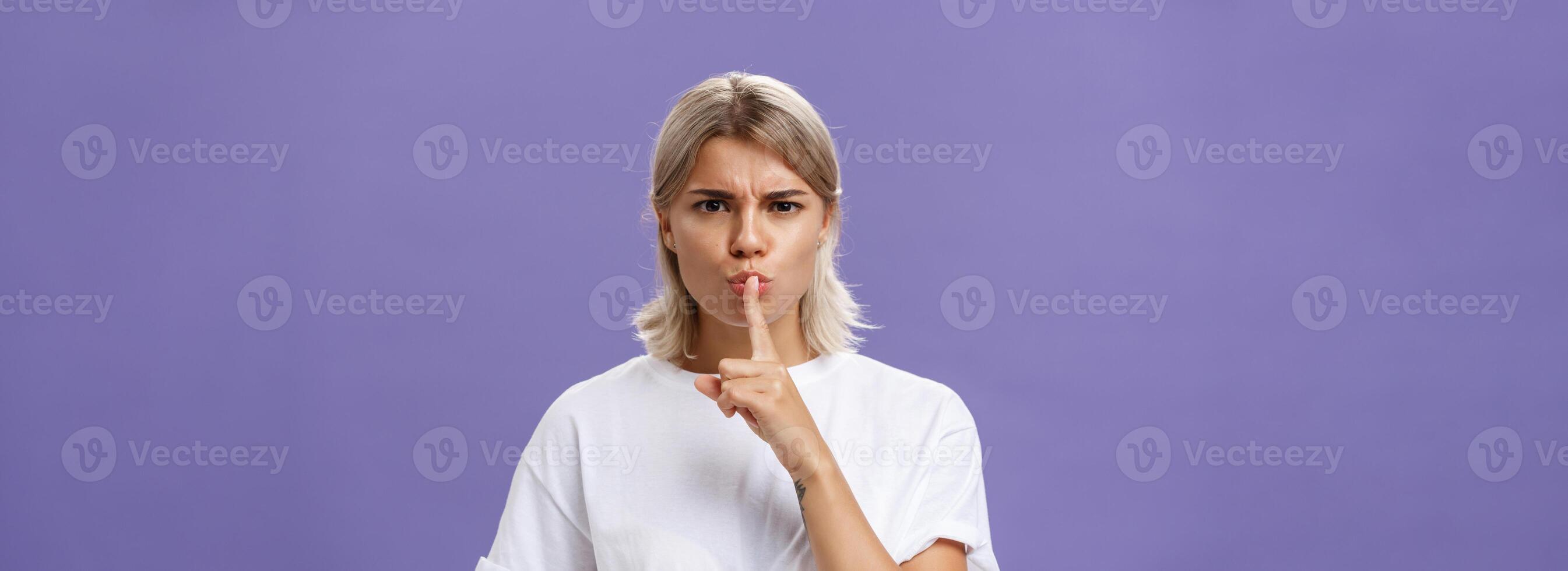 Close-up shot of serious strict good-looking caucasian female with blond hair shushing at camera displeased and disappointed frowning holding index finger over folded lips in shush sign photo