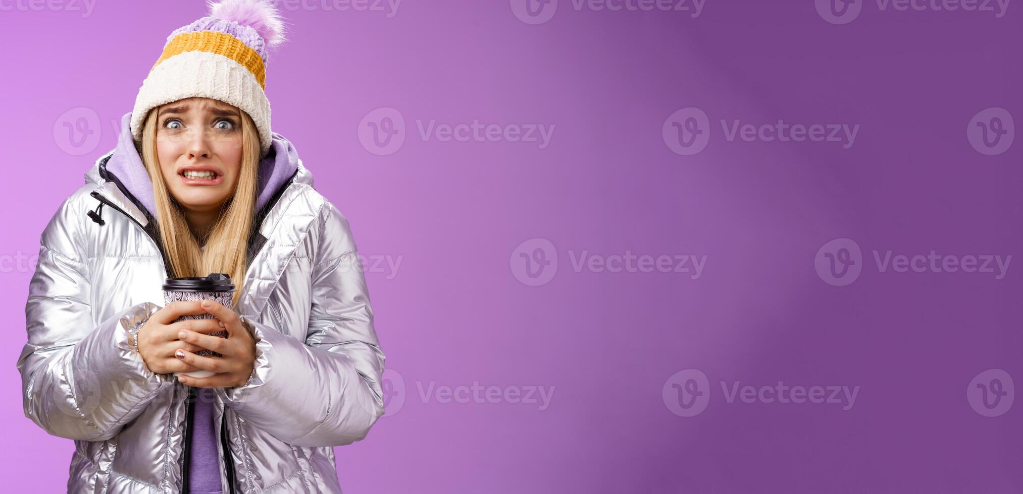 Shaking uncomfortable displeased girl wearing stylish silver jacket hat trembling freezing cold warm hands holding take-away coffee cup clench teeth pop eyes discomfort, standing purple background photo