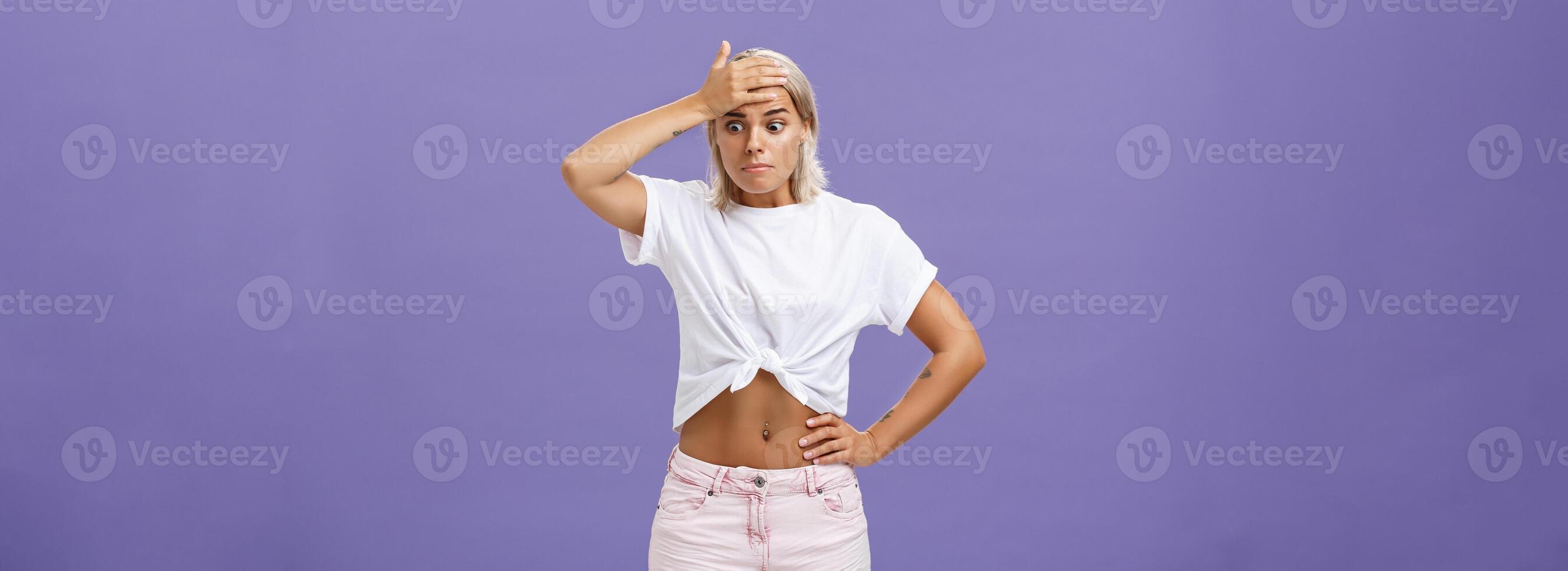 Woman got in serious bad sitation feeling concerned and nervous starting panic holding hand on forehead pursing lips staring down with shocked, perplexed expression being in trouble over purple wall photo