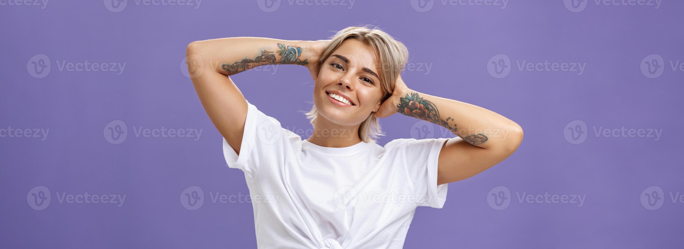 Close-up shot of good-looking stylish and relaxed blond woman with tattoos on arms holding hands behind head smiling with carefree pleased look enjoying weekends over purple background photo