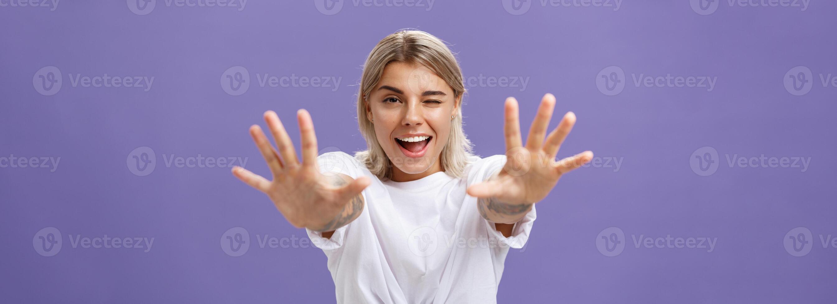 alcanzando a tú. retrato de coqueta guapo y confidente elegante mujer con tatuajes en brazos parpadeo pega fuera lengua en broma y sonriente tracción manos hacia cámara terminado púrpura antecedentes foto