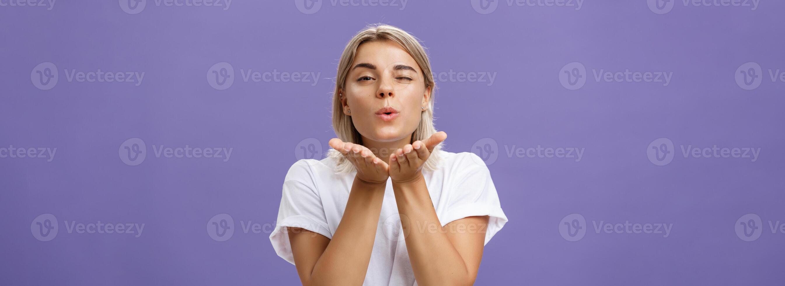 Blowing sweet kiss to all fans. Flirty tender and cute stylish european female with blond hair and tanned skin holding palms near mouth folding lips while sending mwah at camera over purple wall photo