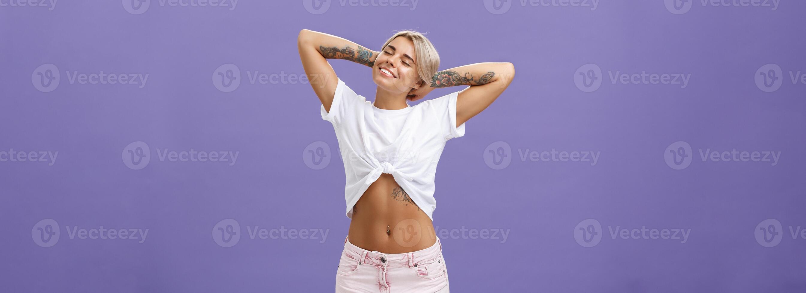 Great feelings after sunbathing. Chill and relaxed attractive young female in cropped top with perfect body and tattoos holding hands behind head, stretching with delight and smiling with closed eyes photo