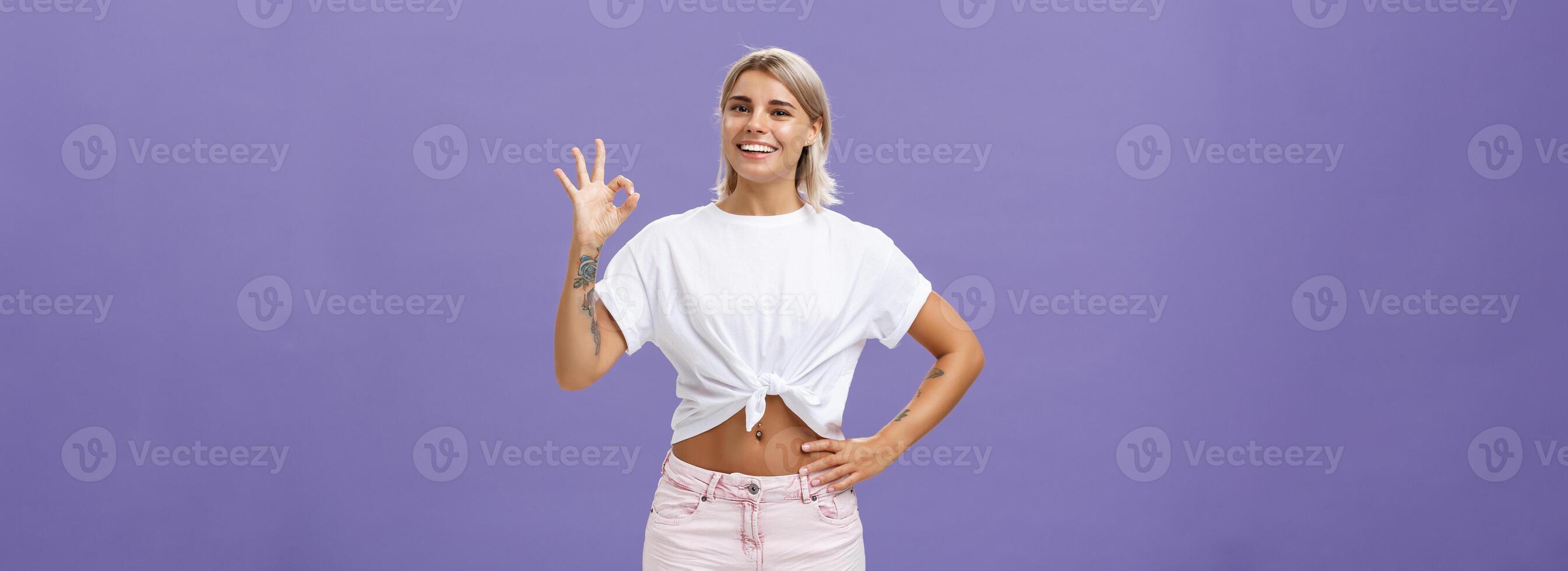 Excellent I like it. Portrait of satisfied good-looking happy girl with fair hair in white t-shirt and shorts showing ok or perfect gesture and smiling broadly holding hand on waist over purple wall photo